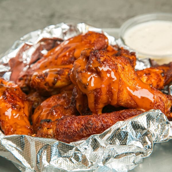 Lenzini's Pizza's buffalo chicken wings, glistening with sauce, served in a foil-lined container with ranch.
