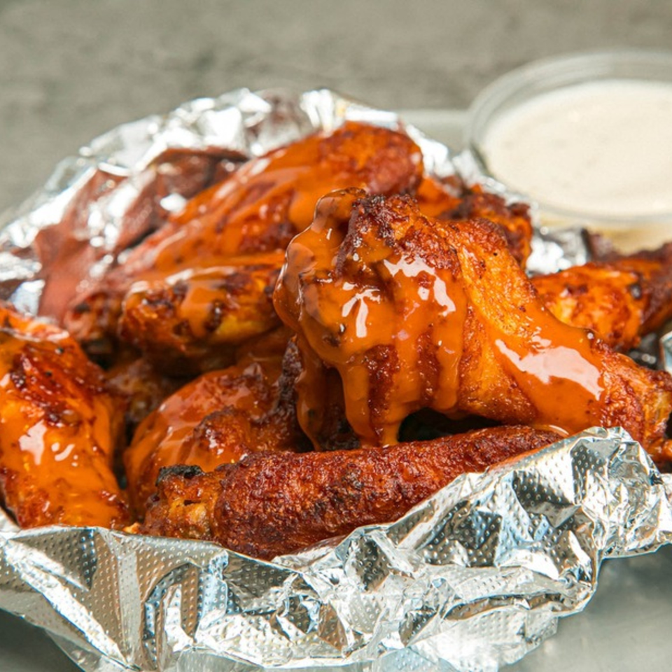 Lenzini's Pizza's buffalo chicken wings, glistening with sauce, served in a foil-lined container with ranch.